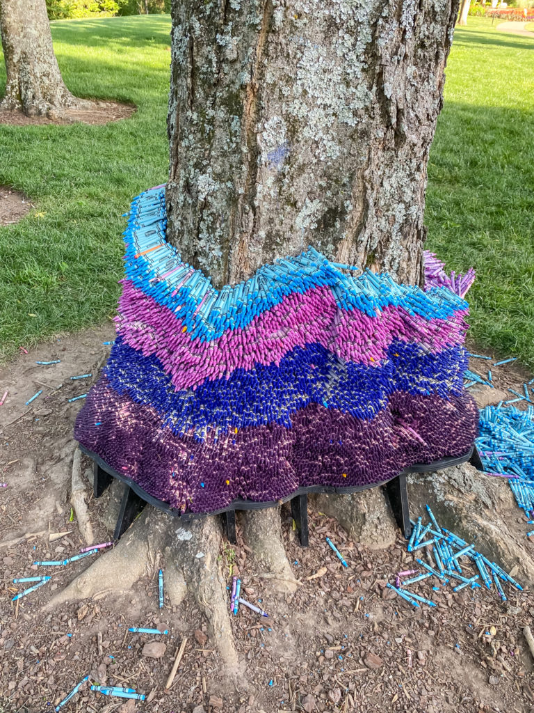 Crayons stacked in a ring around a tree trunk with bottom layer purple, then blue, then light purple, then light blue with some crayons on the ground around the tree. Created by Herb Williams