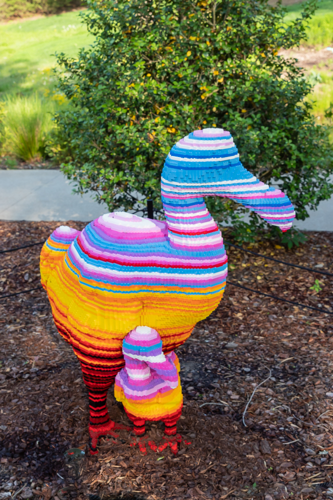 Lego dodo bird sculpture with young dodo bird at its feet. Legs are alternating stripes in shades of red. Lower half of body is yellow and and orange stripes. Upper half is blue, purple, pink, and white stripes.