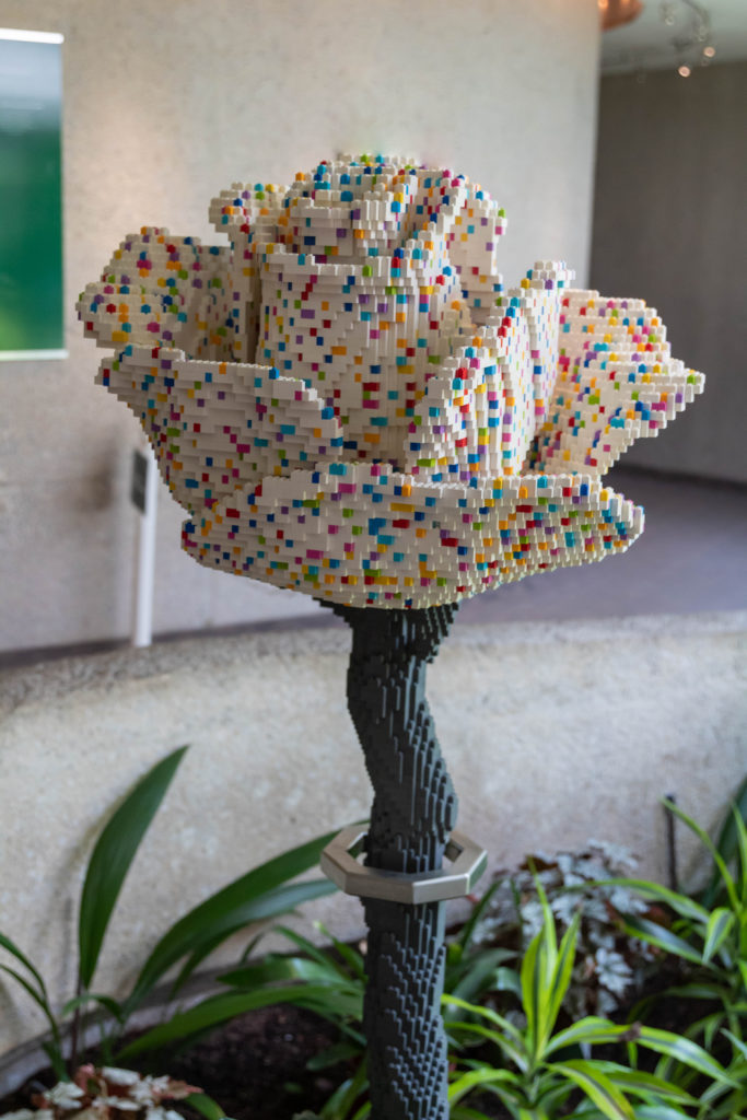 Lego rose with white flower speckled with a rainbow of color blocks. Stem is dark grey in the visitors center of Cheekwood